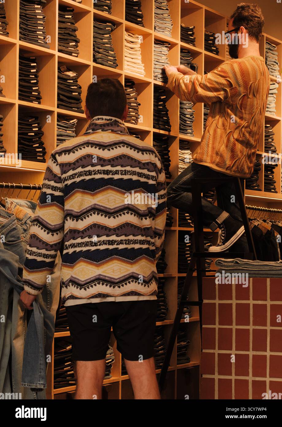 Servizio negozio di jeans: Due uomini, uno su una scala, lavorano in un negozio di abbigliamento caldo e terroso con jeans impilati. Atmosfera al dettaglio realistica e mirata. Foto Stock