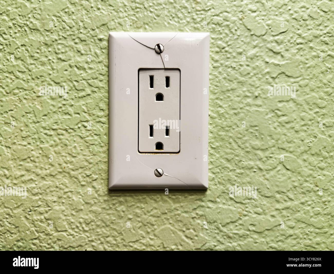 Presa elettrica a muro incrinata con piastra frontale in plastica danneggiata su parete interna verde testurizzata, manutenzione domestica e concetto di sicurezza elettrica Foto Stock