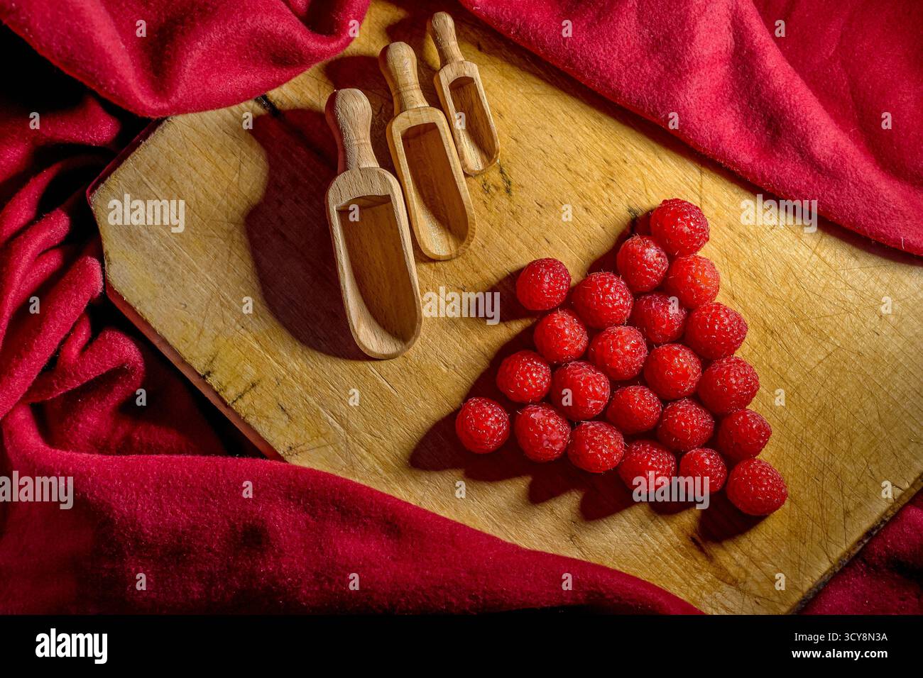 Lamponi freschi disposti a triangolo accanto ai misurini di legno. Lo sfondo in tessuto rosso aggiunge calore e armonia visiva. Foto Stock