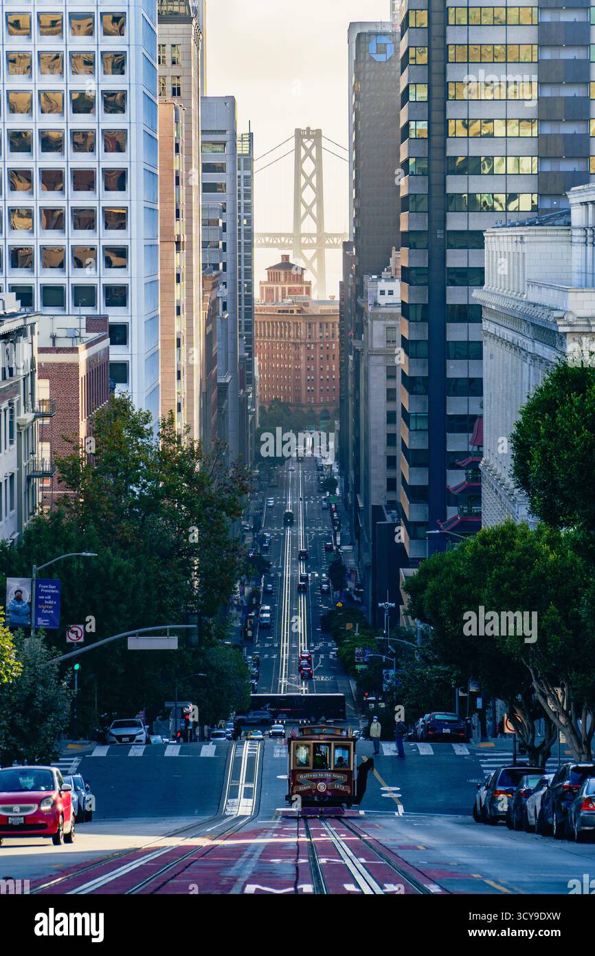 Una classica funivia di San Francisco sale su una ripida strada della città fiancheggiata da un'architettura diversa, con il Bay Bridge visibile sullo sfondo. Foto Stock