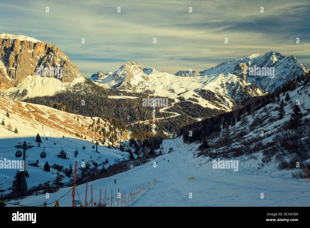 Un'ampia e ben curata pista da sci scende attraverso una valle con gondole e foreste sempreverdi, adagiata contro le massicce cime innevate delle Dolomiti Foto Stock