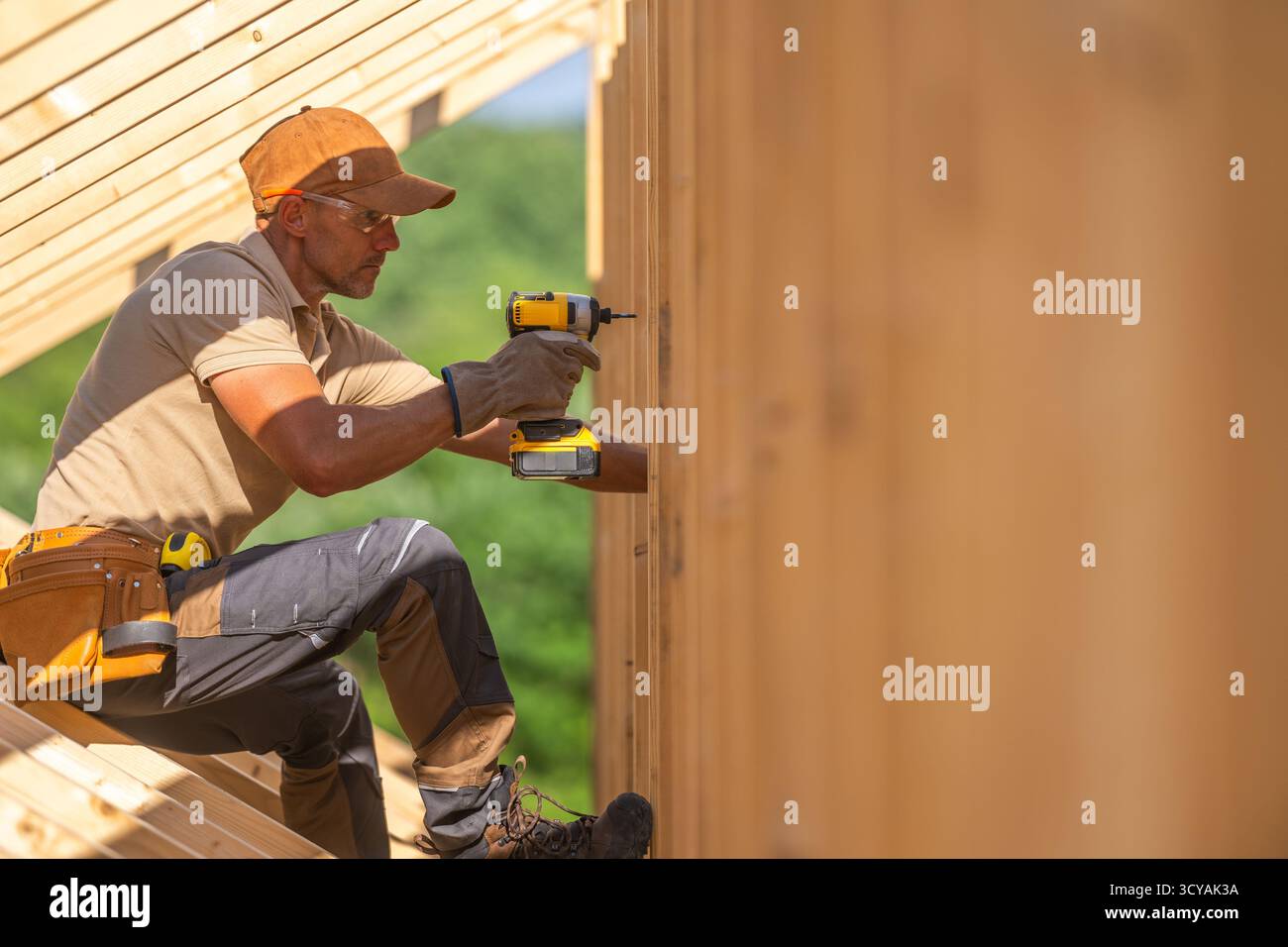 Un lavoratore si concentra sull'assemblaggio di travi in legno, utilizzando un utensile elettrico in un ambiente luminoso e all'aperto. Foto Stock
