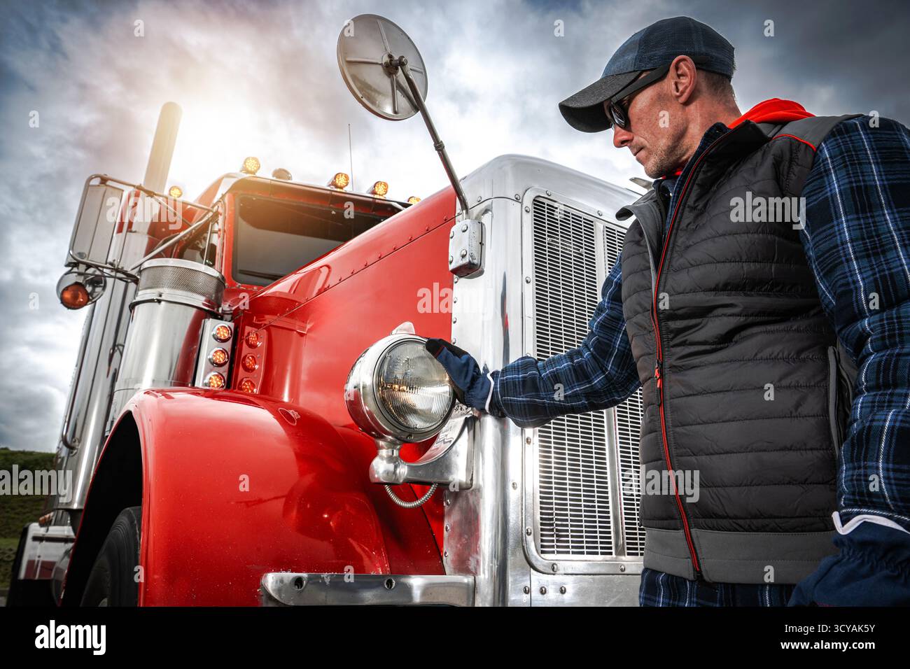 Un uomo con giubbotto e cappellino ispeziona un camion rosso sotto un cielo coperto nel tardo pomeriggio, mostrando i dettagli del veicolo. Foto Stock