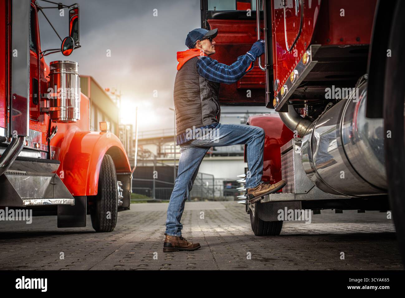 Un conducente di camion sale sul suo veicolo mentre il sole tramonta, pronto per il viaggio che precede. Nelle vicinanze, un altro camion aspetta. Foto Stock