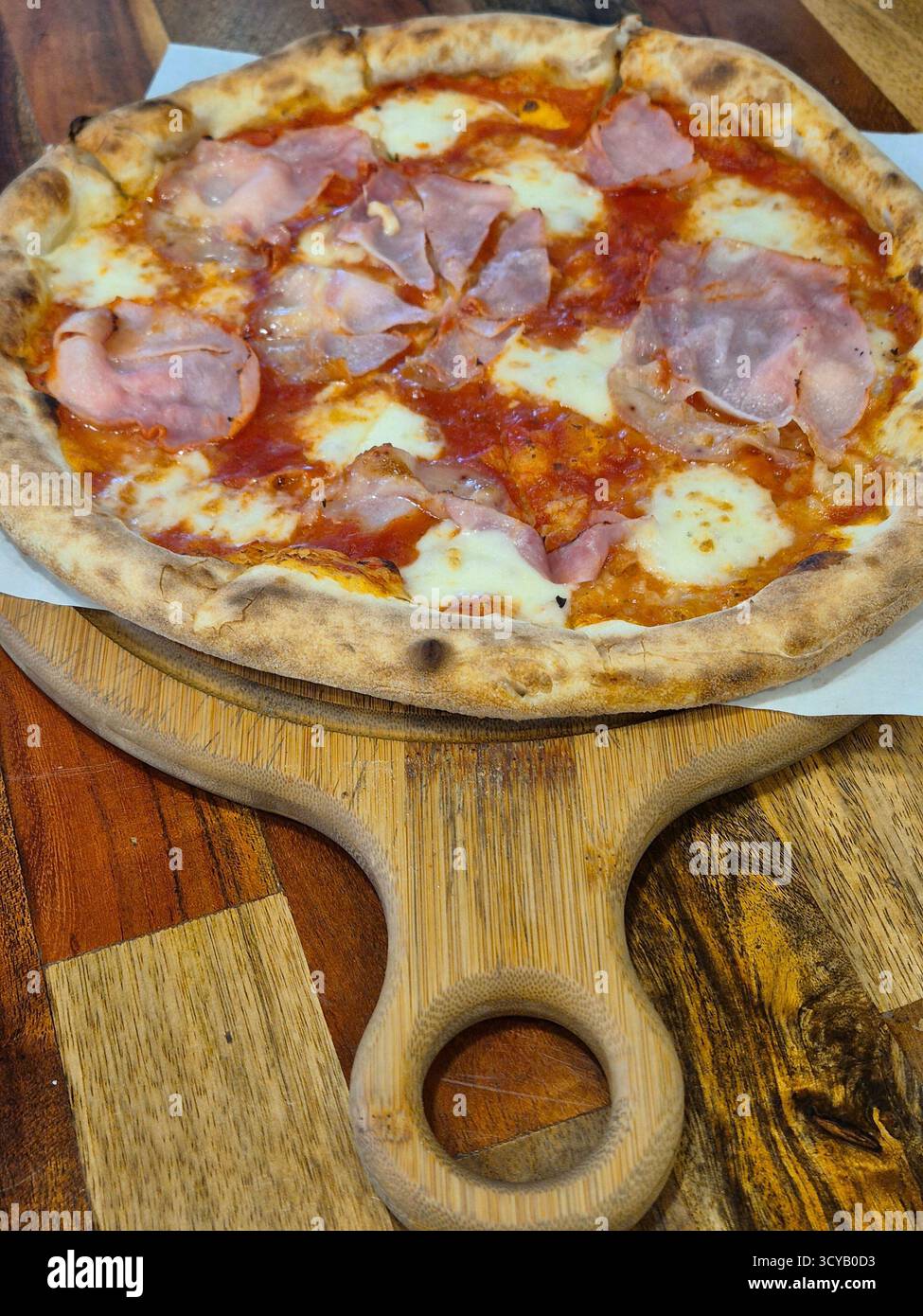 La foto mostra un primo piano di una pizza appena sfornata con prosciutto e formaggio, servita su una tavola rotonda di legno con manico - Immagine stock catturata con smartphone