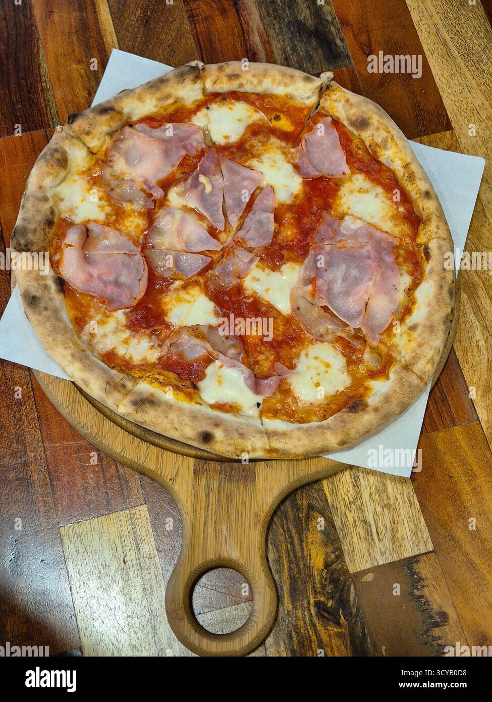 La foto mostra un primo piano di una pizza appena sfornata con prosciutto e formaggio, servita su una tavola rotonda di legno con manico - Immagine stock catturata con smartphone