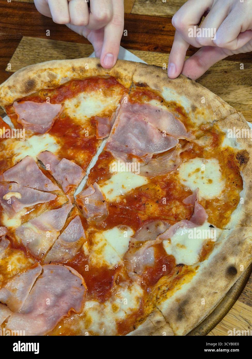 La foto mostra un primo piano di una pizza appena sfornata con prosciutto e formaggio, servita su una tavola rotonda di legno con manico - Immagine stock catturata con smartphone