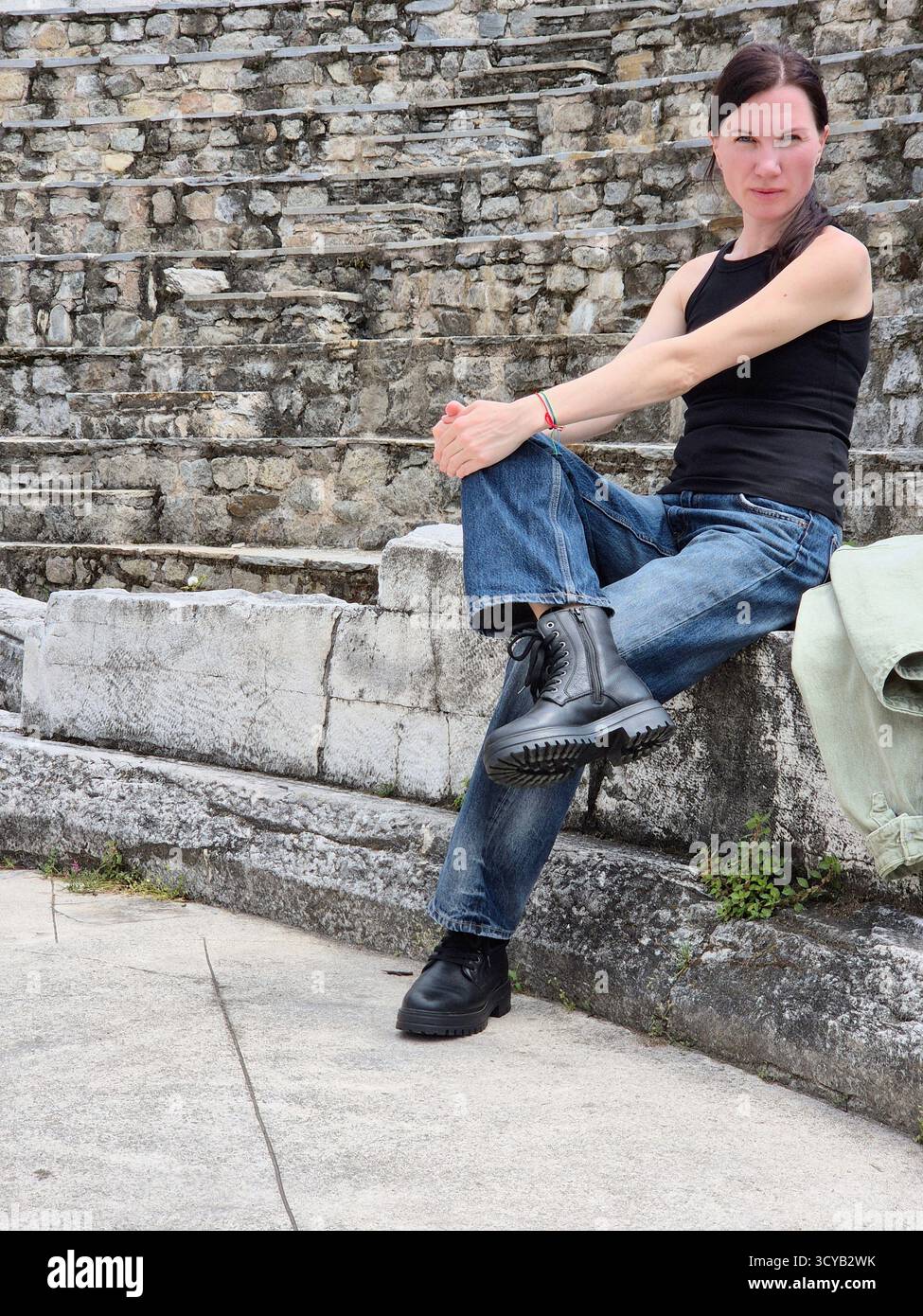 Una donna posa in abiti casual contro un massiccio muro di pietra, sottolineando l'atmosfera di una città antica - Immagine stock catturata con smartphone