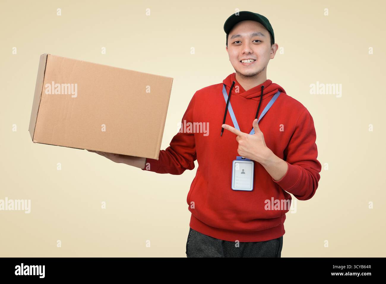 Un corriere sorridente in una felpa con cappuccio e cappuccio rosso può contenere e puntare la confezione sullo sfondo dello studio Foto Stock