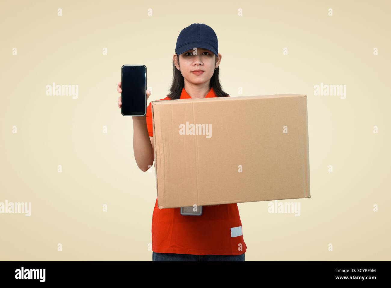 consegna giovane lavoratore con espositore per smartphone e confezione, corriere sorridente pronto per la consegna a casa in studio Foto Stock