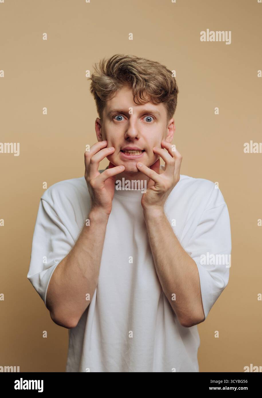 Un giovane si trova in uno studio su uno sfondo chiaro, mostrando paura attraverso gli occhi larghi, la bocca aperta e le mani sul viso. La sua espressione è Foto Stock