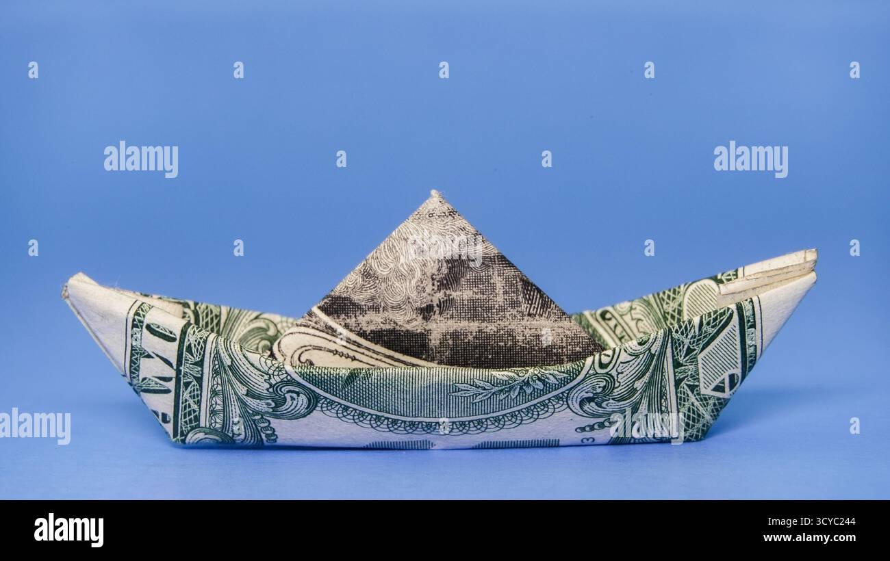 Una banconota da un dollaro è abilmente piegata per formare una barca seduta su un morbido sfondo blu. Questa creazione unica mette in mostra sia l'arte che la finanza in un gioco Foto Stock