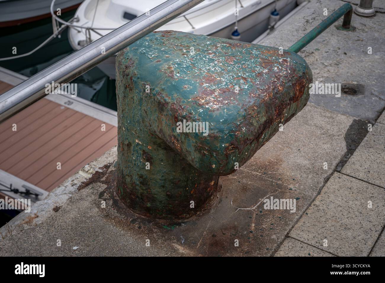 Dissuasore in metallo color verde acqua, fortemente arrugginito, imbullonato a un molo in cemento in un tranquillo porticciolo, che mostra segni di deterioramento e deterioramento Foto Stock