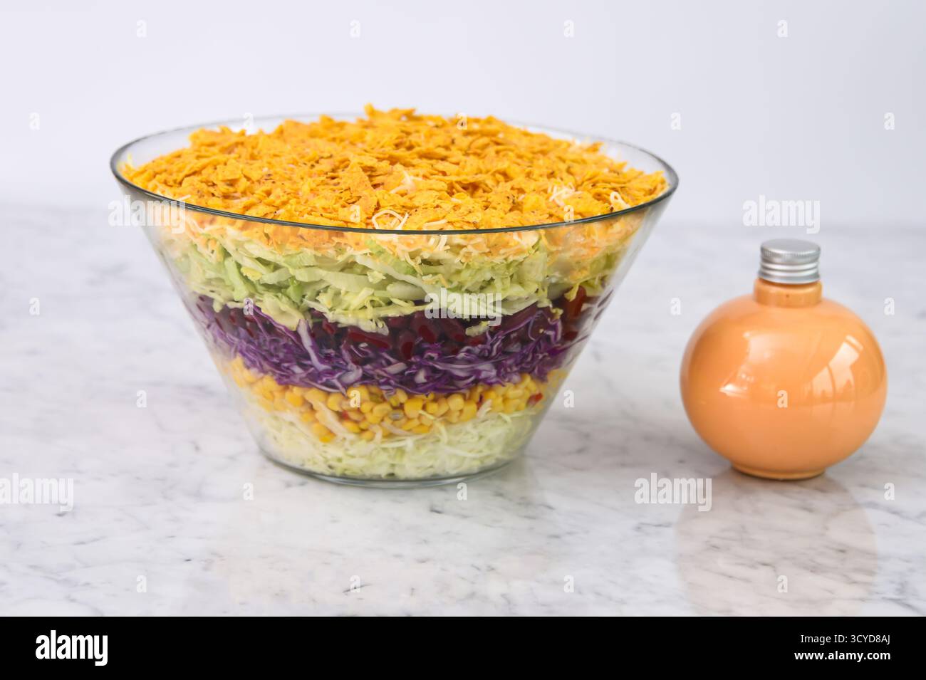 Insalata a sette strati in un recipiente di vetro trasparente con strati di lattuga grattugiata, mais, cavolo rosso, cavolo verde, bottiglia di salsa cremosa, e un croccante schiacciato Foto Stock