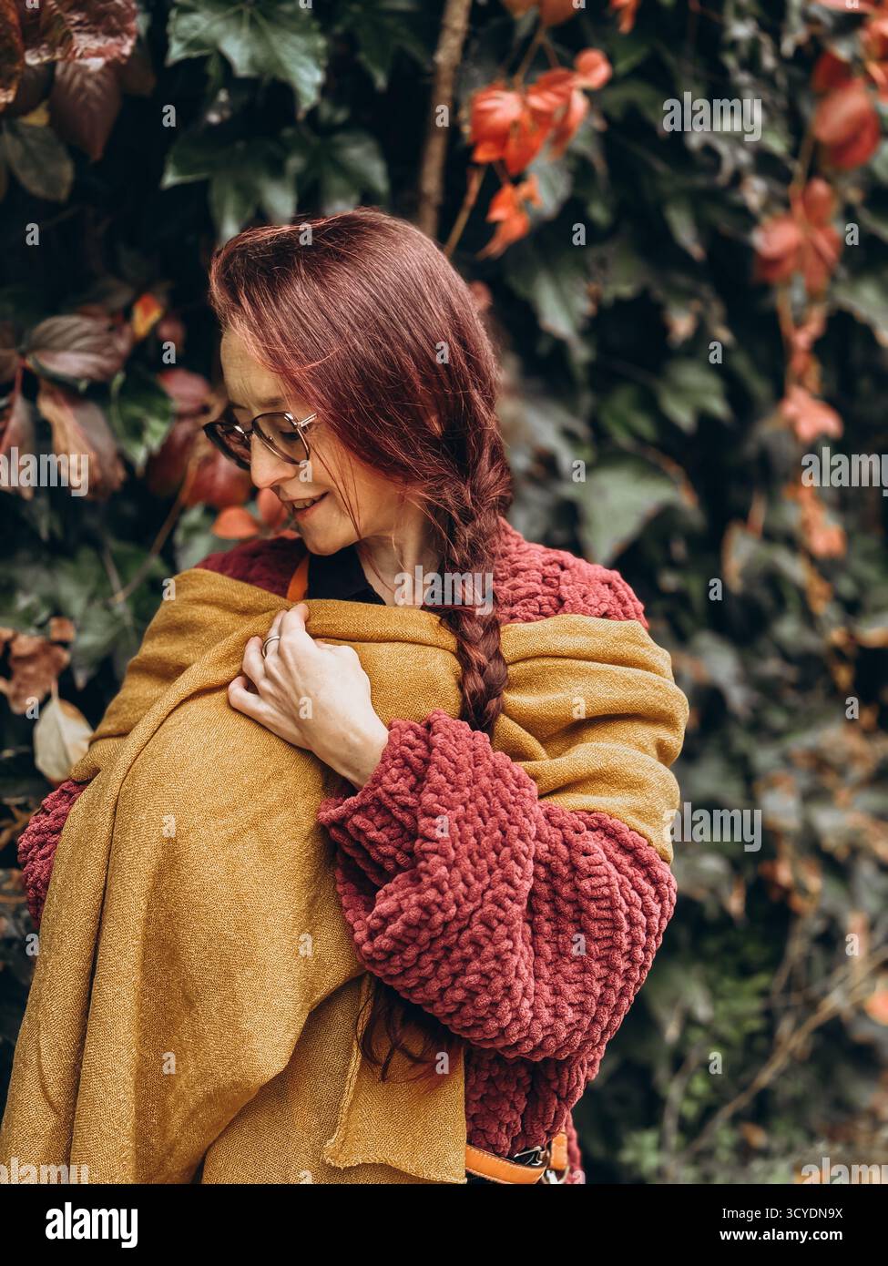 Donna con sciarpa gialla senape e maglione testurizzato rosso con capelli intrecciati in piedi vicino alla parete edera morbido umore autunnale accogliente stile di vita lento tranquillo Foto Stock