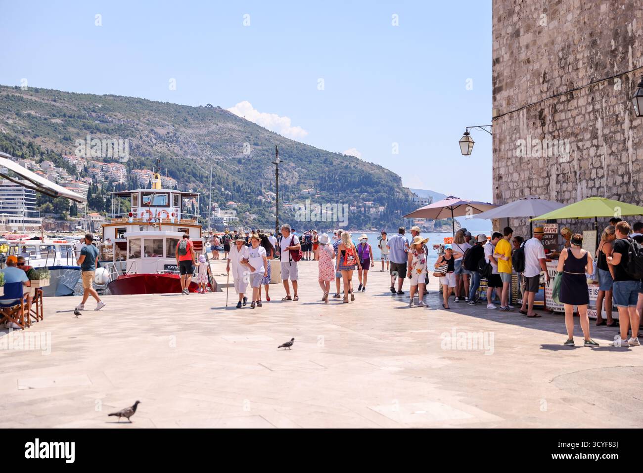 I turisti si allineano per le escursioni in barca lungo il porto vecchio di Dubrovnik, Croazia, con montagne e navi da crociera visibili sullo sfondo. Foto Stock