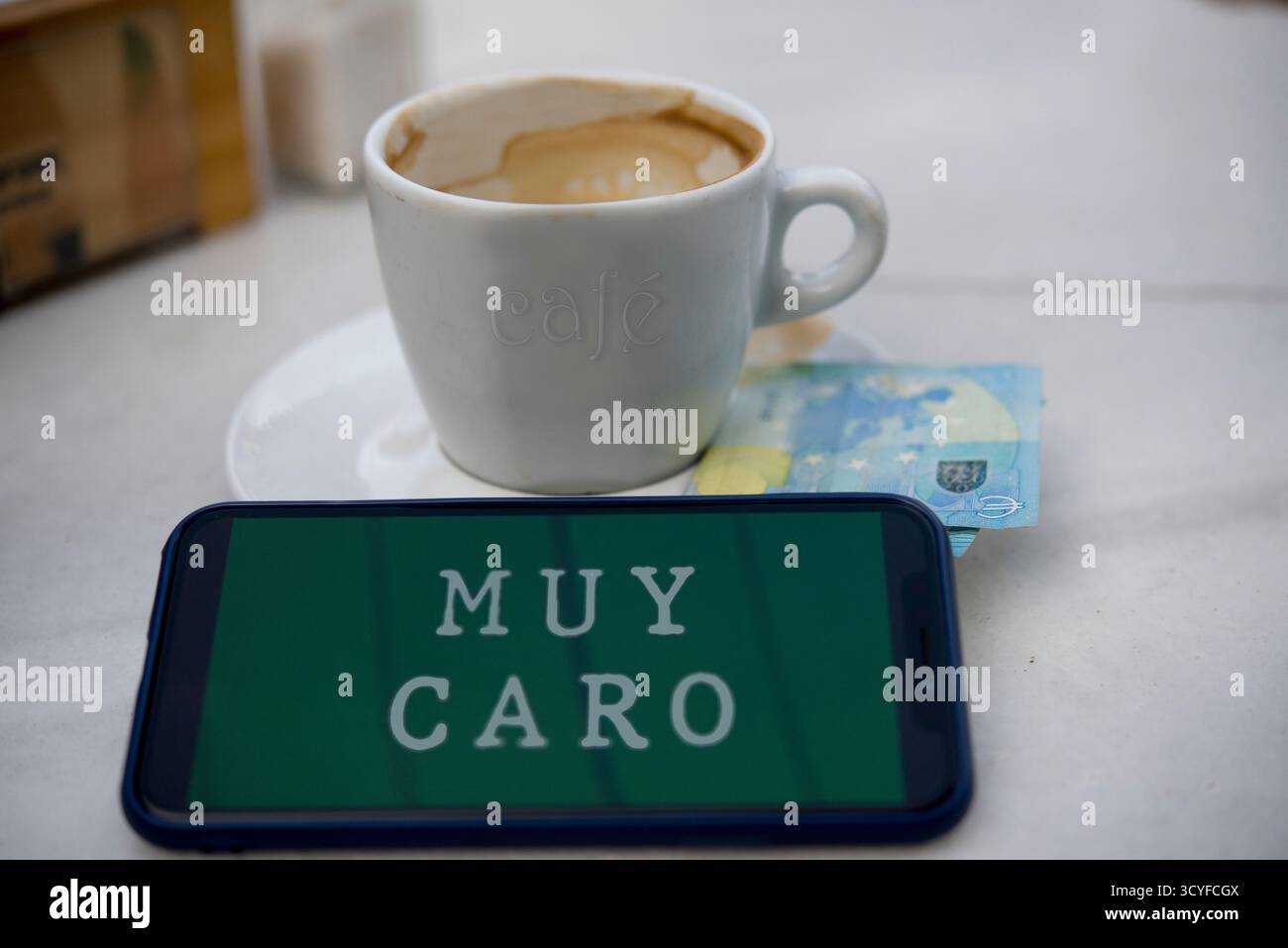 Una tazza di caffè vuota con un telefono che dice "troppo costoso" accanto a una banconota da 20 euro su un piatto, che fa riferimento ai prezzi elevati del caffè e alle abitudini di consumo urbano Foto Stock