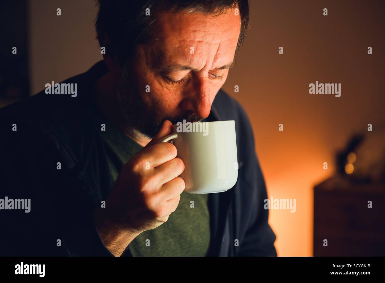Maschio beve tè caldo a casa nella calda luce serale, rilassandosi e godendo un momento di tranquillità. Calmo momento introspettivo di un uomo che sorseggia il tè, simboleggiando Foto Stock