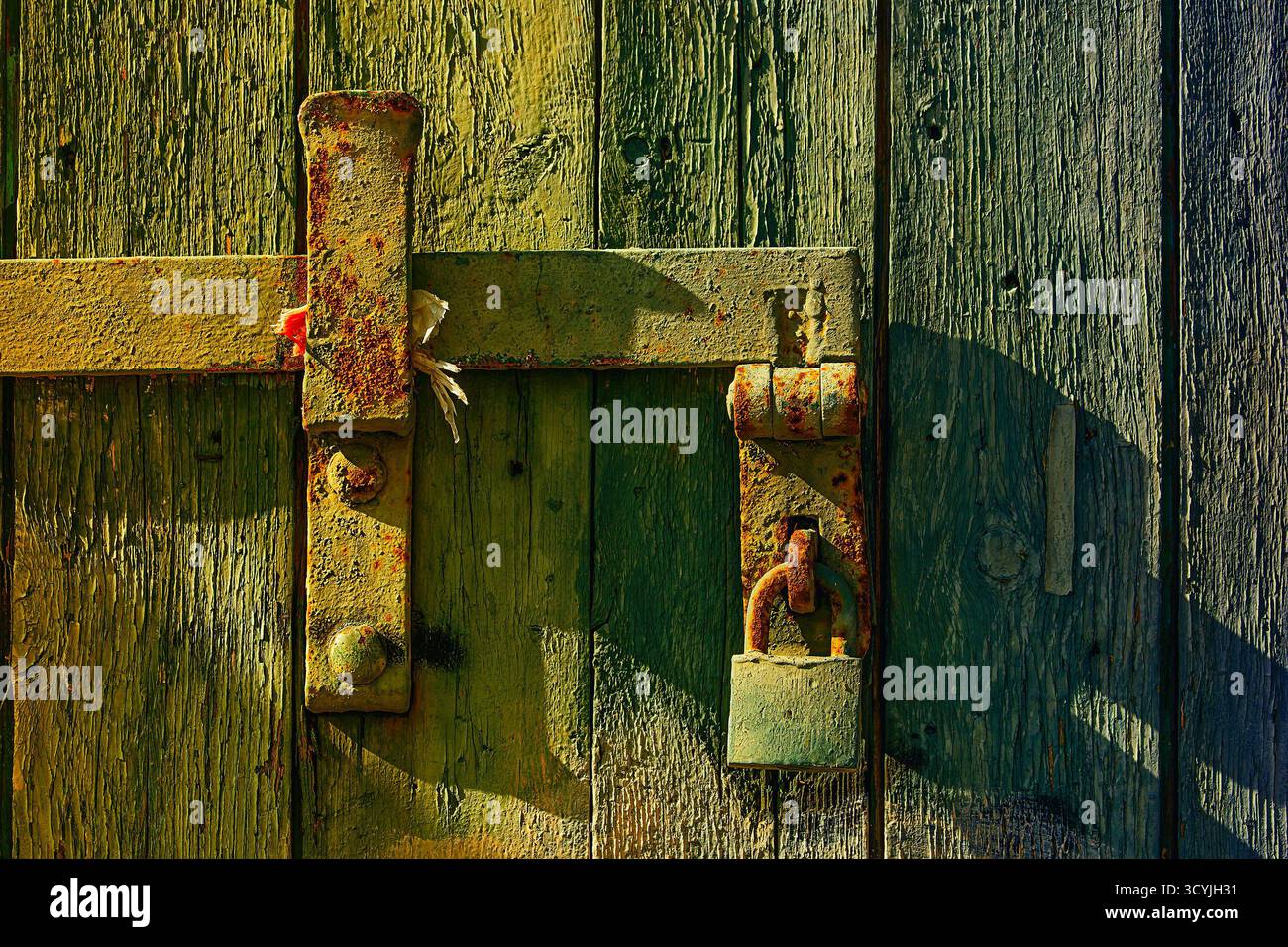 Un primo piano di un vecchio lucchetto in metallo arrugginito su una porta in legno resistente agli agenti atmosferici, inondato dalla calda luce del sole pomeridiano, che esalta la consistenza e i colori invecchiati di Th Foto Stock