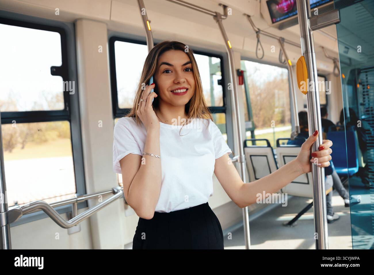 Una donna con i capelli lunghi e una camicia bianca si impegna in una conversazione telefonica mentre si tiene su un palo in un veicolo di transito. La luce del giorno la illumina Foto Stock Una donna con i capelli lunghi e una camicia bianca si impegna in una conversazione telefonica mentre si tiene su un palo in un veicolo di transito. La luce del giorno la illumina Foto Stock