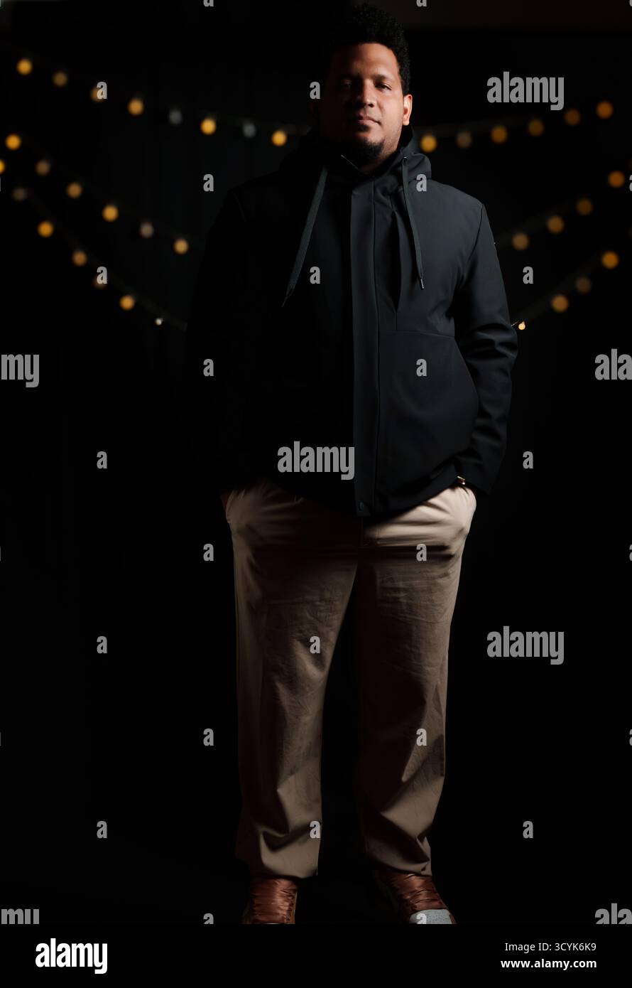 Un uomo con giacca nera e pantaloni tan. E' di fronte a uno sfondo nero Foto Stock