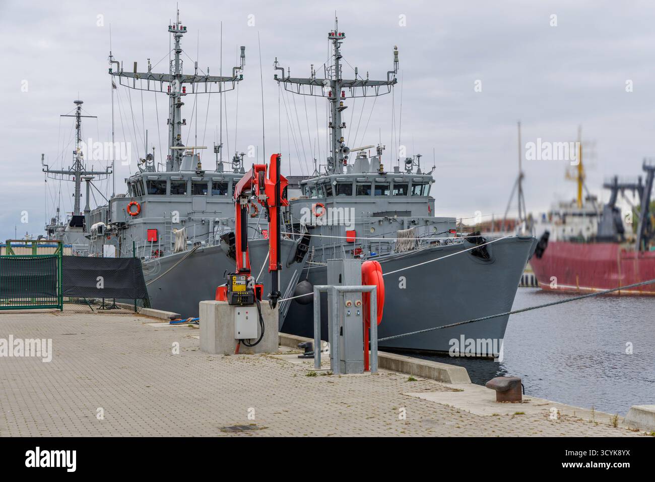 Due navi militari grigie sono ormeggiate vicino a un molo di cemento, caratterizzato da strutture complesse ad albero e una nave rossa offuscata sullo sfondo. Foto Stock