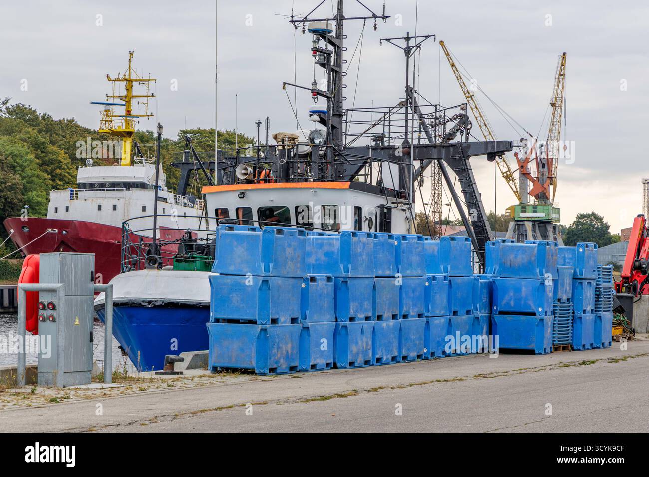 Un'affollata zona portuale industriale con una nave da pesca o da servizio ormeggiata, contenitori di plastica blu impilati in primo piano e gru da carico gialle Foto Stock