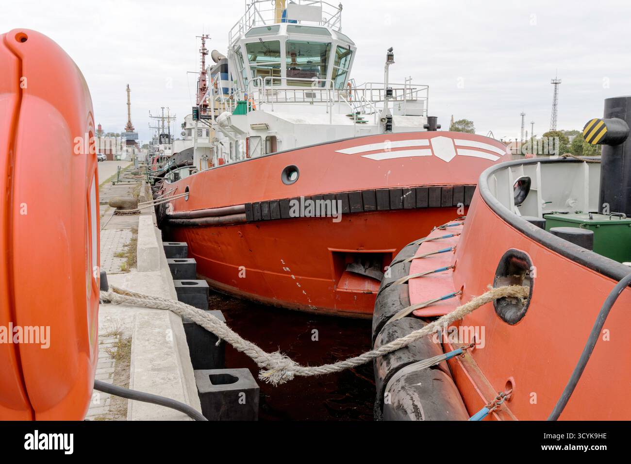 Un'ampia ed ampia vista di un porto industriale che mostri una grande nave da carico o da servizio con scafo rosso ancorata, con container impilati blu, una gru gialla, Foto Stock