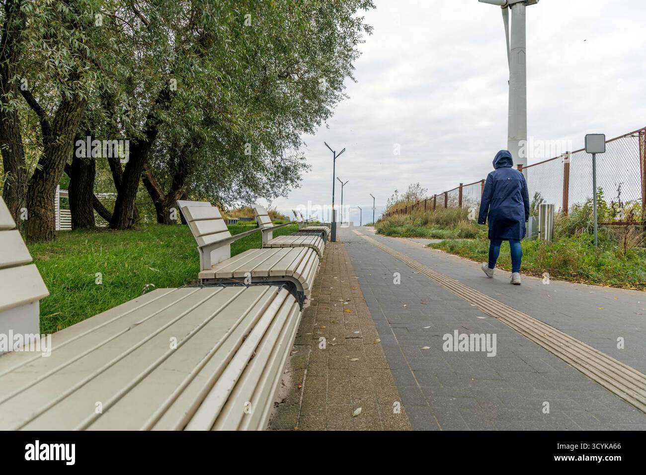 Una persona con una giacca blu con cappuccio si allontana lungo un sentiero lastricato, passando davanti a una fila di panchine moderne dai colori chiari e alberi verdi Foto Stock