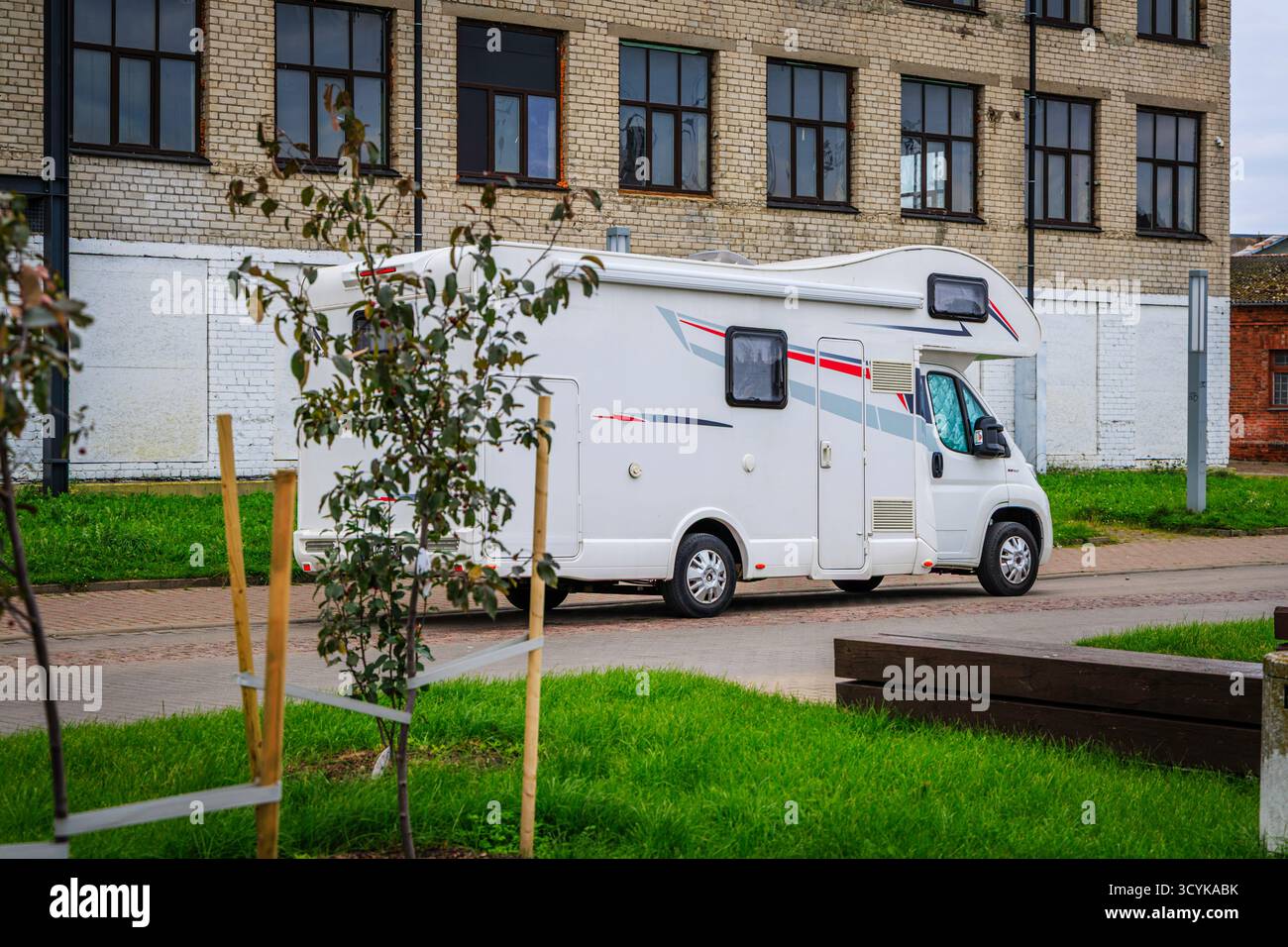 Vista laterale di un camper (veicolo ricreativo) bianco grande e moderno o di un camper parcheggiato in una strada della città vicino a un vecchio edificio industriale piastrellato e in mattoni. Foto Stock