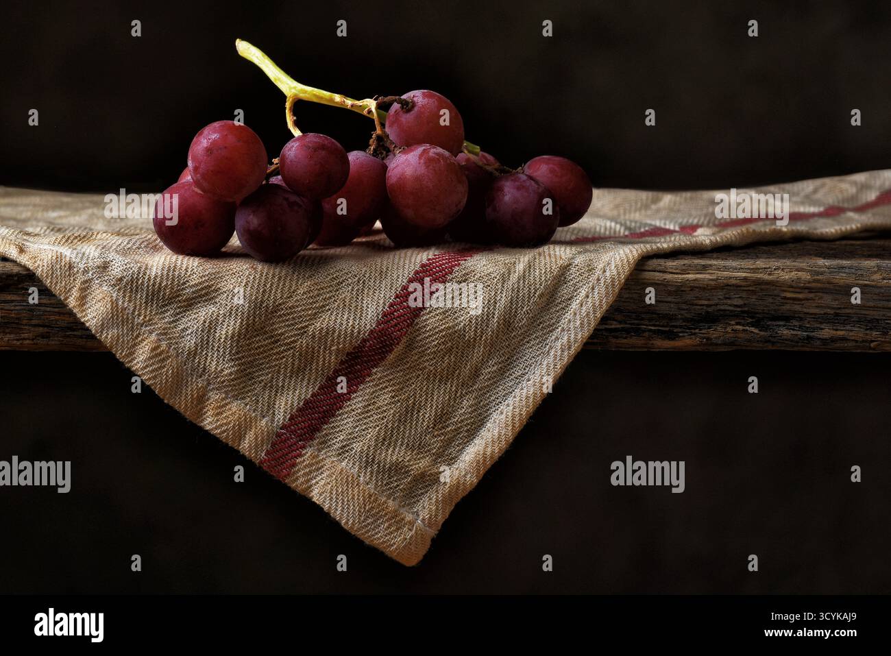 Red Grape natura morta. Un piccolo mazzo di uva rossa matura su un asciugamano da cucina drappeggiato sul bordo di un rustico tavolo di legno con un backgroun marrone strisciato Foto Stock