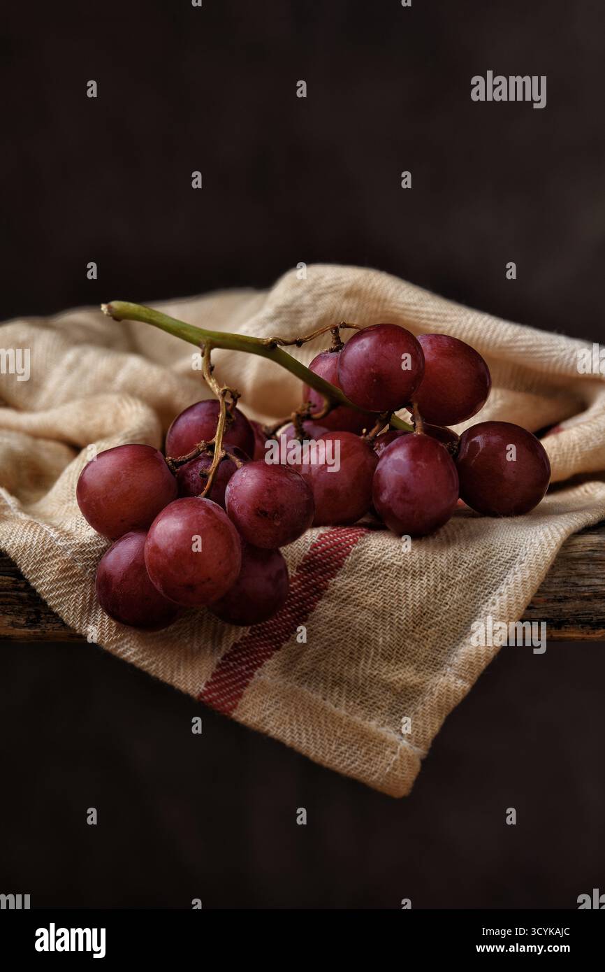 Red Grape natura morta. Primo piano di un mucchio di uva rossa matura su un asciugamano da cucina avvolto sul bordo di un rustico tavolo in legno con un bac marrone a chiazze Foto Stock