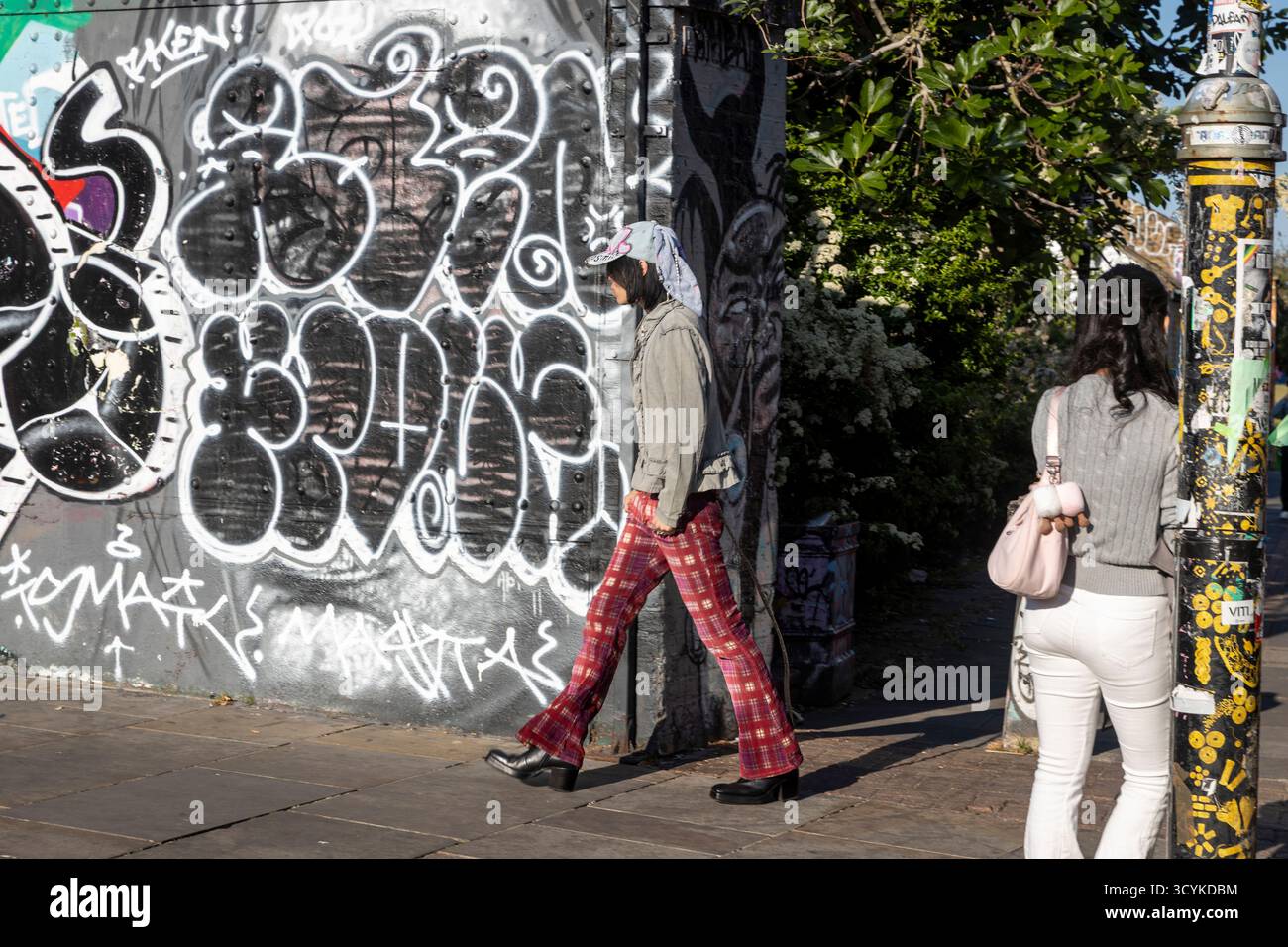 Londra, Regno Unito, 2 maggio 2025. Elegante persona con pantaloni a quadri che passa davanti a un muro di graffiti a Shoreditch Foto Stock