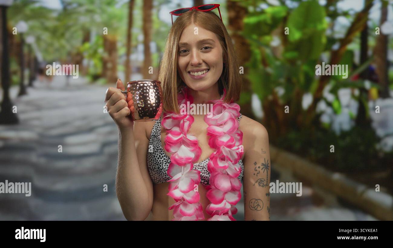 Donna sorridente che tiene in mano una tazza di rame in un vivace parco tropicale con vegetazione lussureggiante che indossa un lei rosa e un costume da bagno elegante, che incarna una spensieratezza Foto Stock