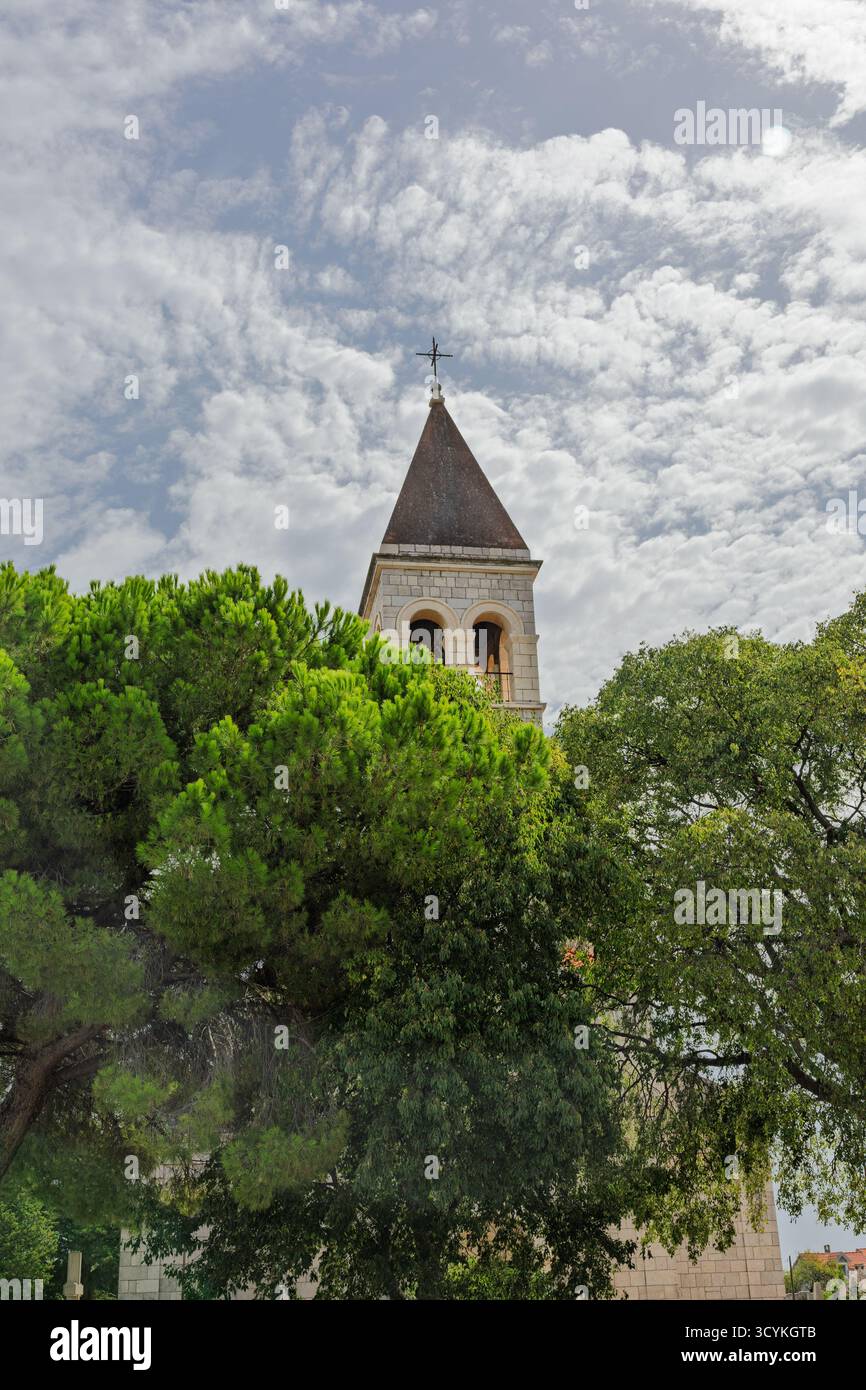 Vecchia chiesa in pietra caratterizzata da un caratteristico campanile, circondata da vegetazione lussureggiante e bagnata dalla luce del sole, su un cielo azzurro limpido e vibrante che raffigura Foto Stock