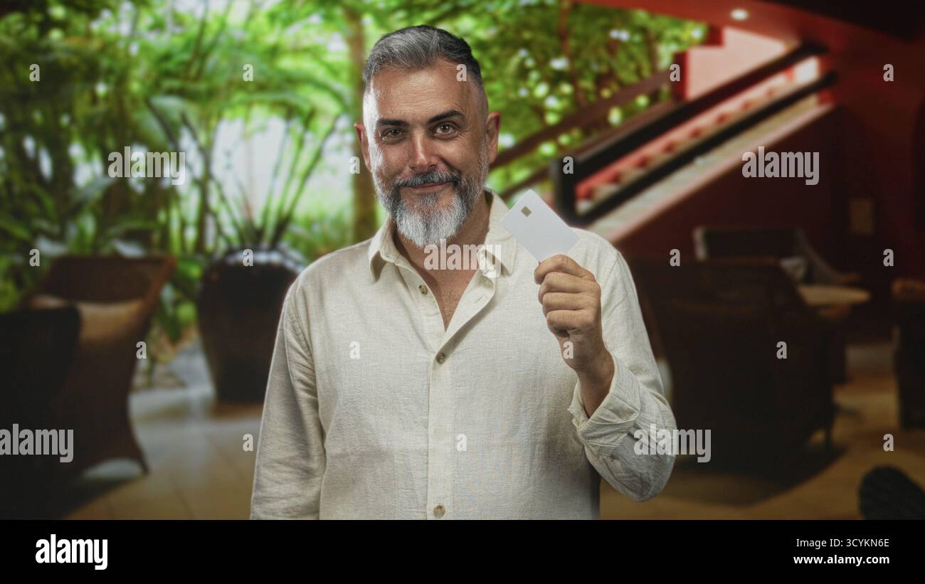 Uomo di mezza età che tiene in mano una chiave bianca nella hall dell'hotel, sorride e strizza l'occhio mentre mostra mano e dita; viaggia per piacere. Foto Stock