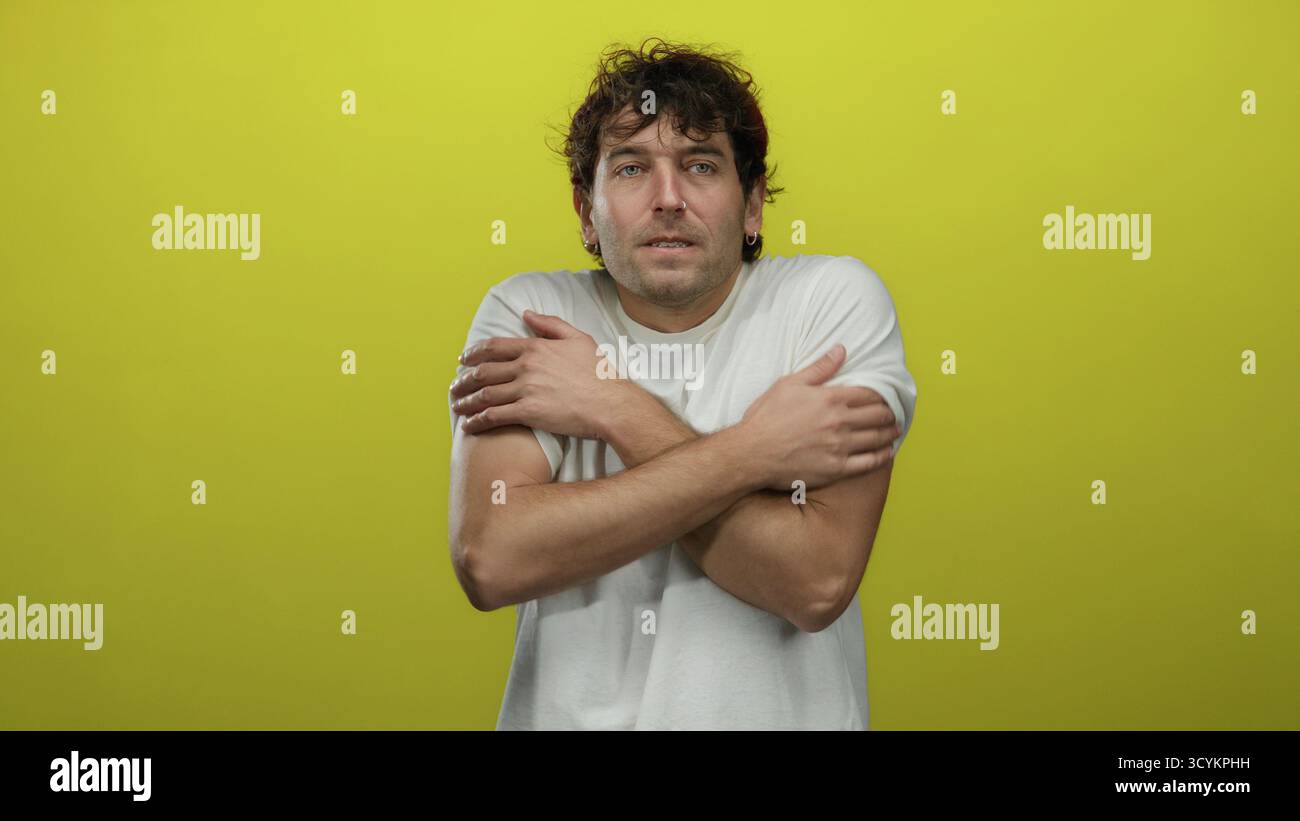Uomo ispanico in camicia bianca che trema dal freddo su sfondo giallo brillante che mette in mostra disagio e ambiente freddo. Foto Stock