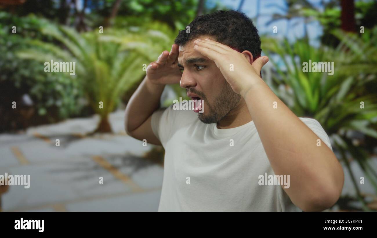 Un giovane che protegge gli occhi con le mani sulla fronte nel passaggio verde del parco sotto la luce del sole e indossa una camicia bianca; sorprendi stupore. Foto Stock