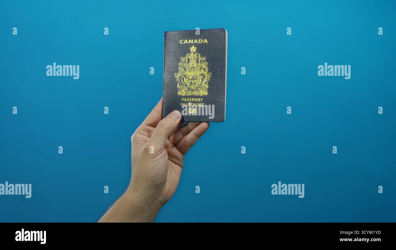 Uomo che ha un passaporto canadese contro un muro blu, che mostra l'identità di viaggio in un ambiente isolato, sottolineando il documento e la mano che tiene Foto Stock