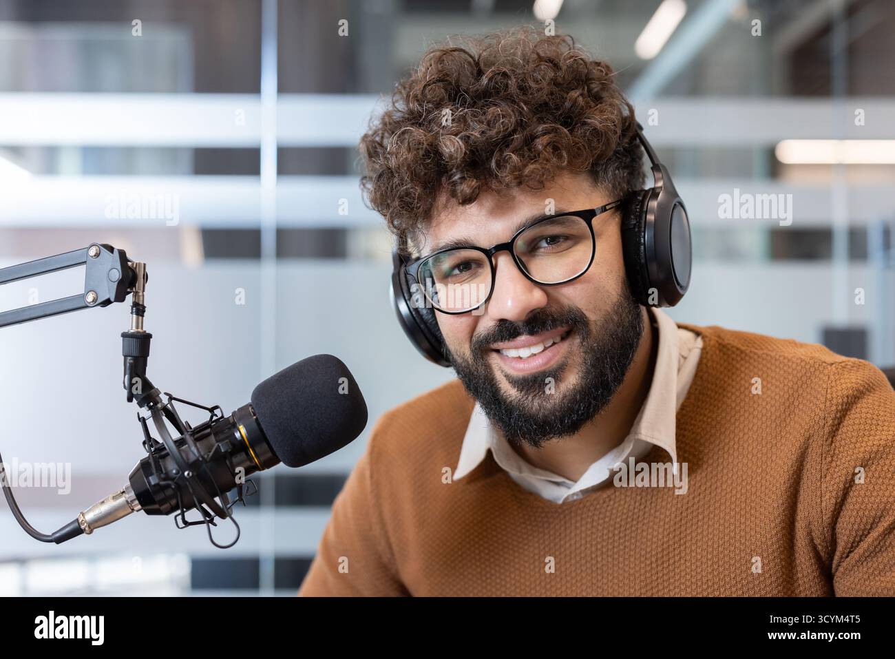 Giovane con capelli ricci e occhiali sorridenti mentre registra un podcast in uno studio moderno, indossa le cuffie e parla in un microfono per contenuti audio online Foto Stock