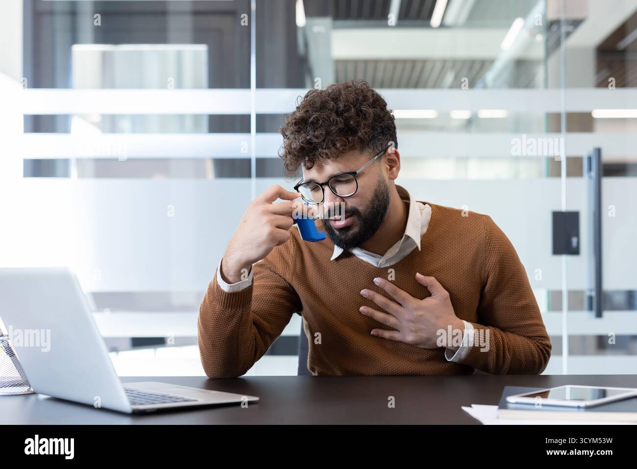 Giovane che soffre di un attacco d'asma e ha difficoltà respiratorie sul posto di lavoro, usando un inalatore blu mentre tiene il petto con preoccupazione Foto Stock