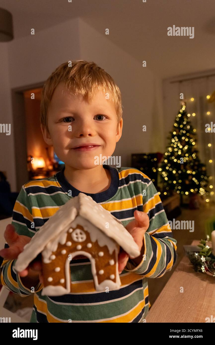 Felice biondo in maglione a righe che mostra orgogliosamente la casa di pan di zenzero fatta in casa con caminetto e luci dell'albero di Natale in un accogliente interno. Vacanze Foto Stock