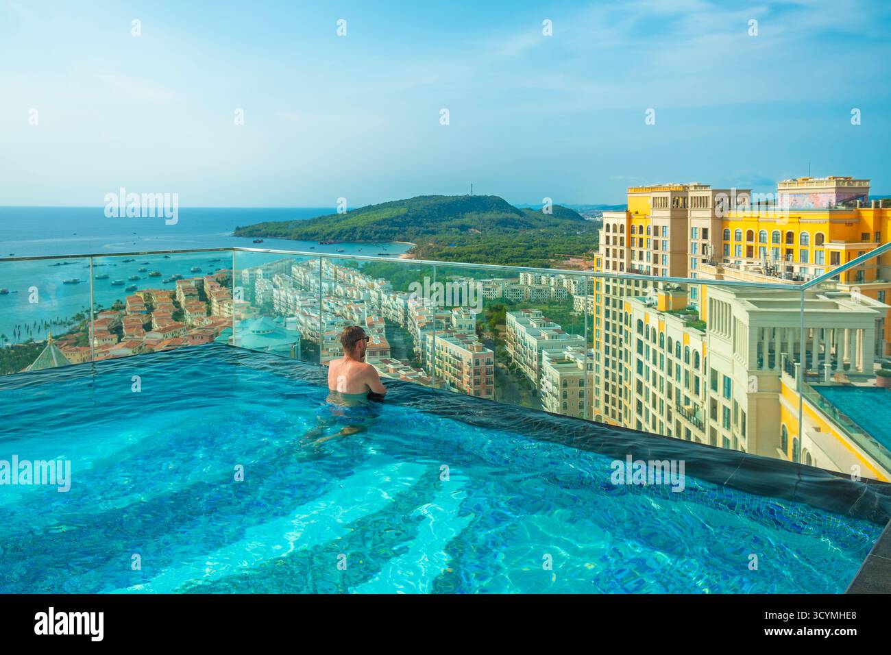 Uomo che si rilassa nella piscina a sfioro sul tetto con splendida vista panoramica dell'isola tropicale e degli edifici in stile mediterraneo nella città Sunset di Phu Quoc Foto Stock