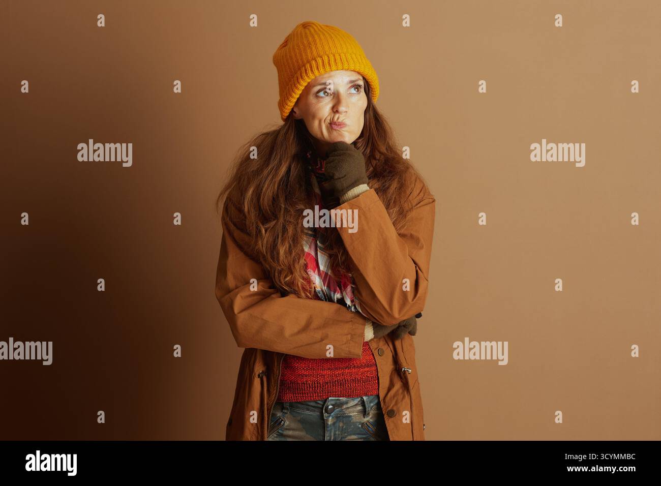 Una donna premurosa con un berretto giallo, un cappotto marrone e un maglione rosso poggia il mento sulla mano, guardando verso l'alto con un'espressione incerta contro un pla Foto Stock