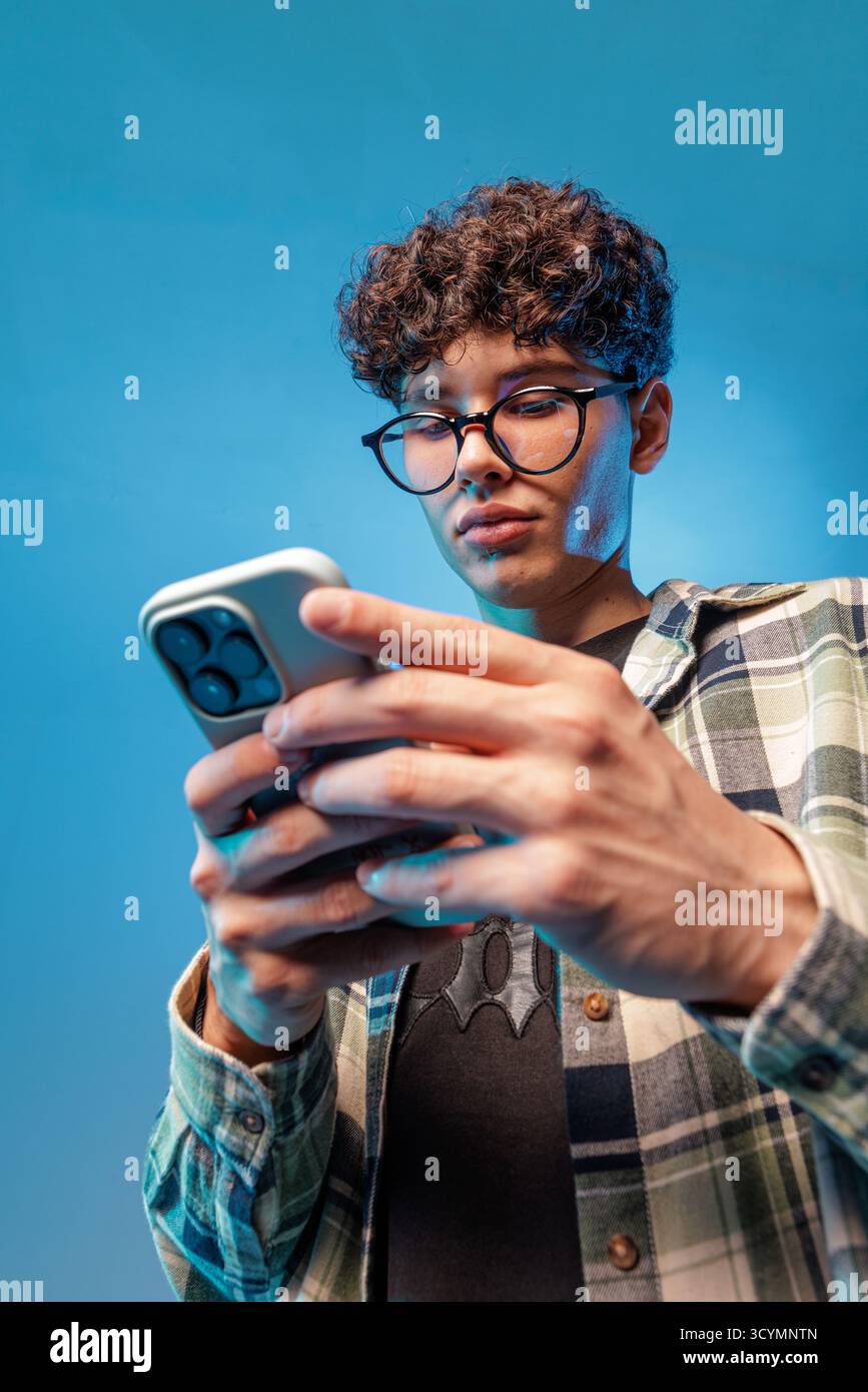 Teenager concentrato con occhiali utilizzando uno smartphone in studio, che illustra l'ora dello schermo, la luce blu, i messaggi di testo e le moderne abitudini digitali. Foto Stock