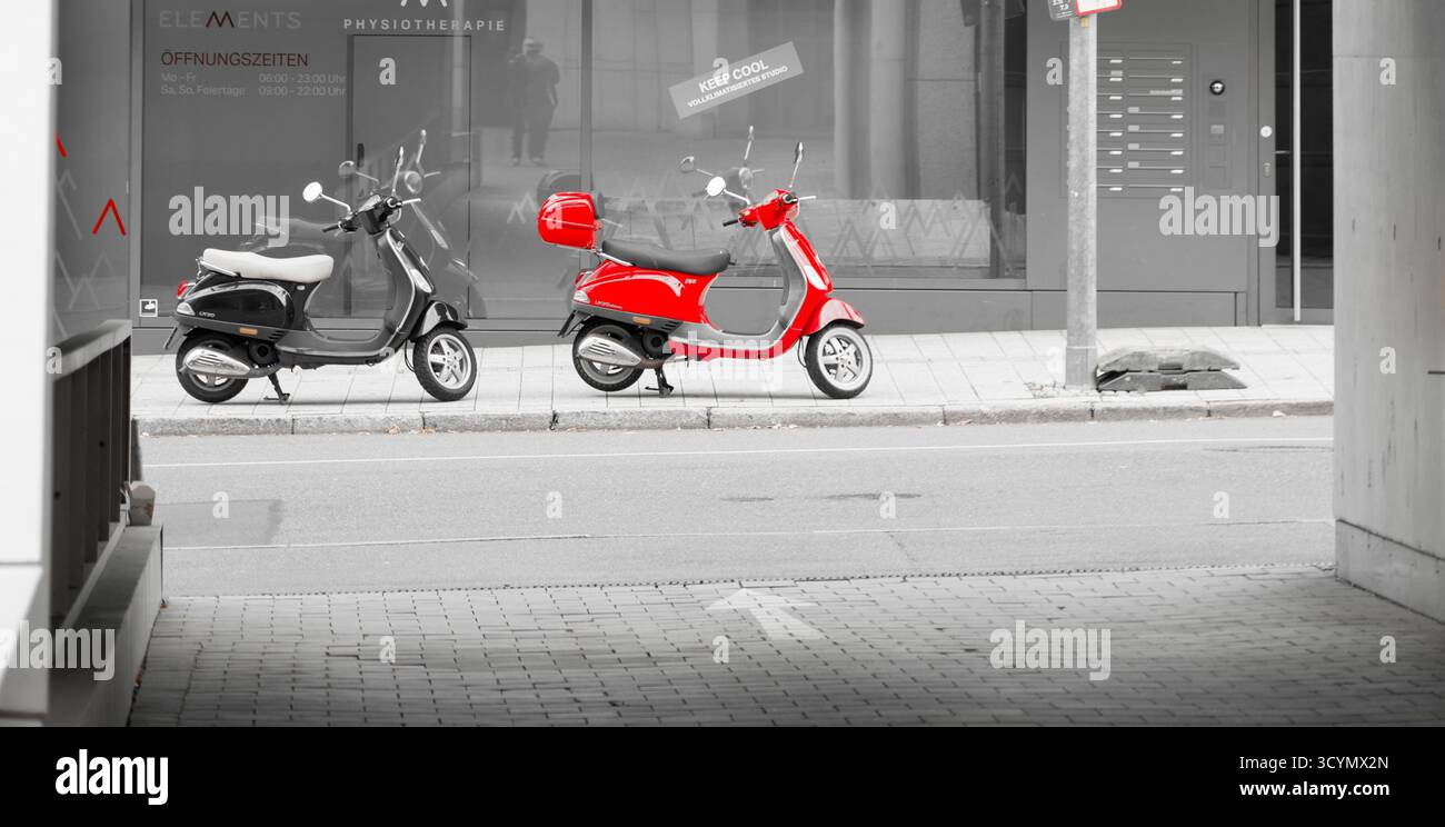 Stoccarda, Germania, 10.18.2025; foto chiave a colori che mostra un ciclomotore rosso su una strada della città, vista attraverso un cancello, con l'ambiente in nero e bianco Foto Stock