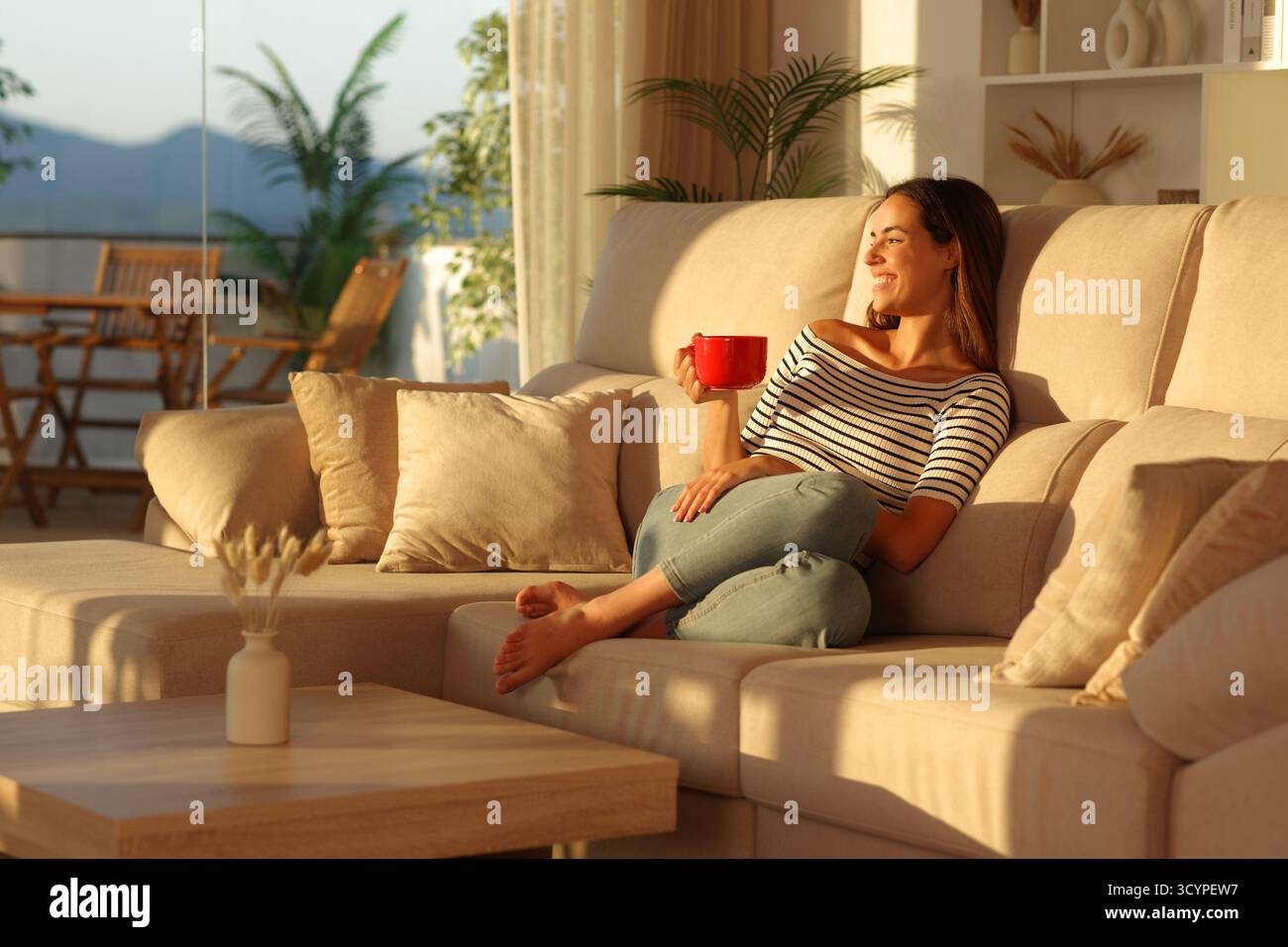 Donna felice che guarda attraverso una finestra bevendo un caffè da una tazza rossa all'interno di una casa al tramonto Foto Stock