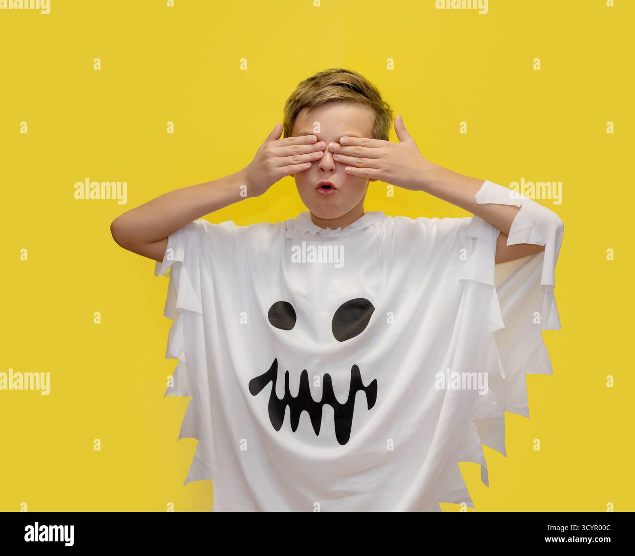 Il ragazzo sorridente vestito da fantasma punta verso l'alto, in piedi di fronte a un vivace sfondo giallo, probabilmente a tema per Halloween. Boy in Ghost Costume poi Foto Stock