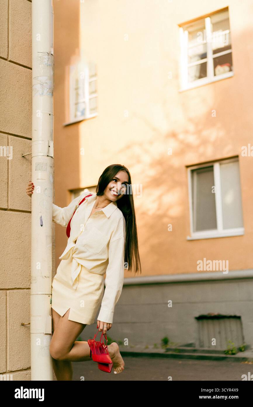 Giovane donna che posa giocosamente all'aperto, tiene in mano una borsa rossa e sorride Foto Stock