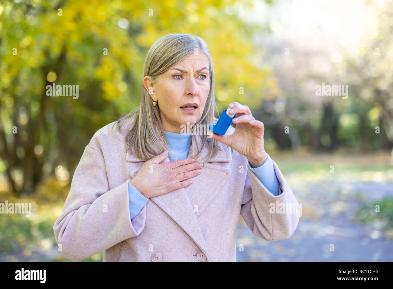 Donna anziana con un'espressione ansiosa che sta vivendo un attacco d'asma in un parco, tenendo in mano un inalatore e ponendo una mano sul petto, cercando sollievo dai problemi respiratori Foto Stock